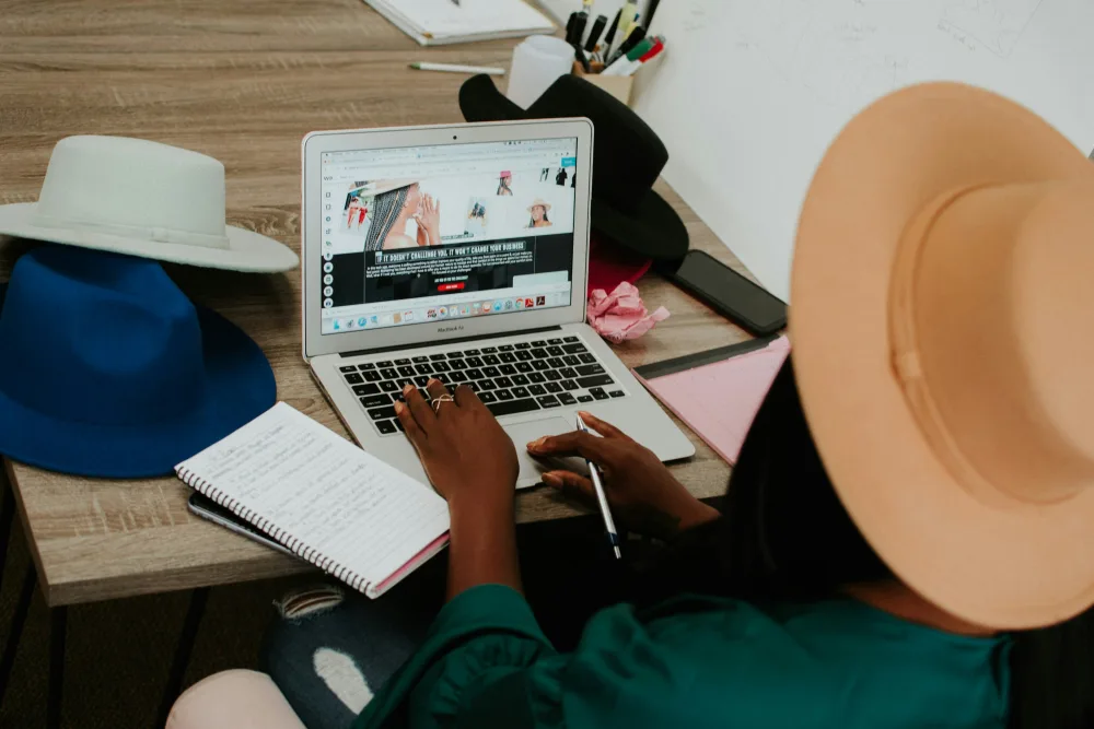A milliner works at a laptop in her workshop, surrounded by hats.