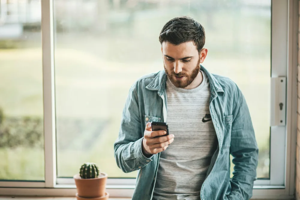 A man in a denim shirt reads a message on his phone by a window.