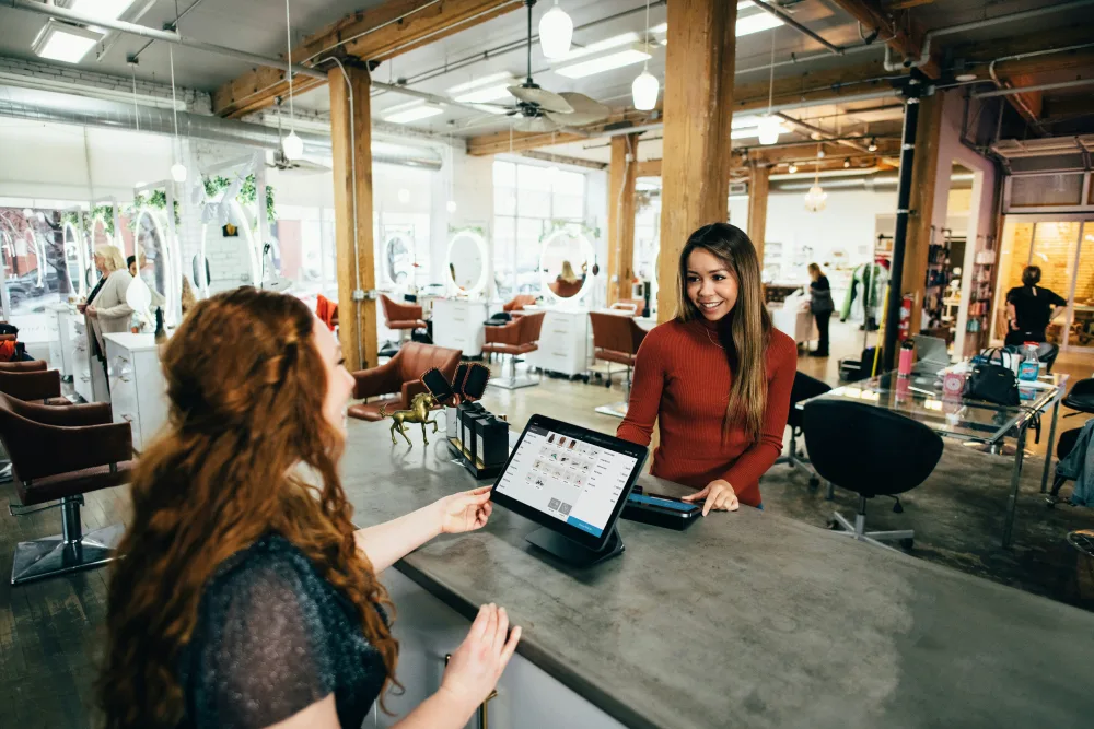 A salon owner serves a customer at a tablet till point in a busy salon.